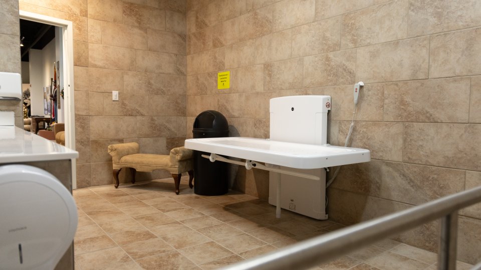 Spacious beige-tiled restroom with a wall-mounted adult changing table. A plush chair and black trash can are near the door, creating a functional, welcoming space.