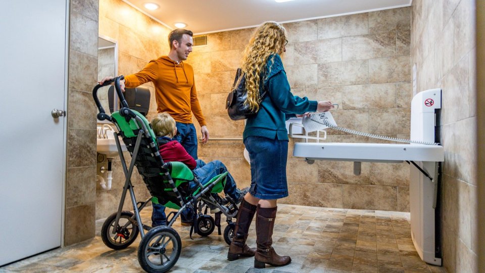 A man in an orange hoodie and a woman with long hair stand in a tiled room with a stroller. The woman adjusts a height-adjustable changing table.