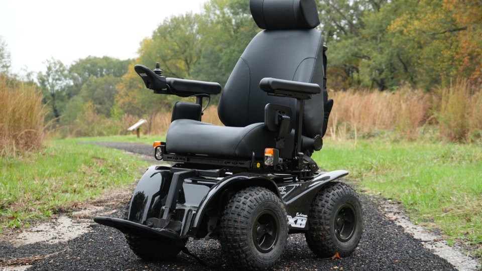 A rugged black power wheelchair sits on a winding gravel path in a lush park with tall grass and trees, conveying adventure and independence.