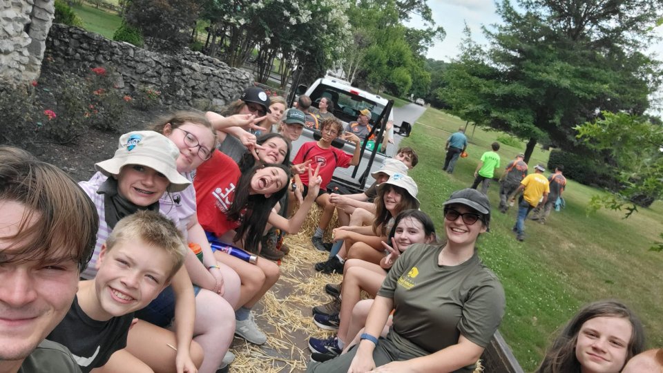 A group of smiling teenagers sits on hay bales in a trailer, driven by a white truck. They appear cheerful and are making peace signs. Trees and grass are in the background, conveying a fun, outdoor setting.