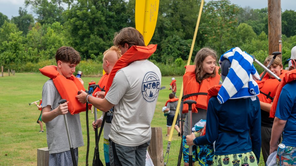 A group of kids wearing orange life vests gather outdoors for rafting. One holds a yellow paddle, while another's head is wrapped in a blue towel. The setting is a lush, green park, creating a lively, adventurous atmosphere.