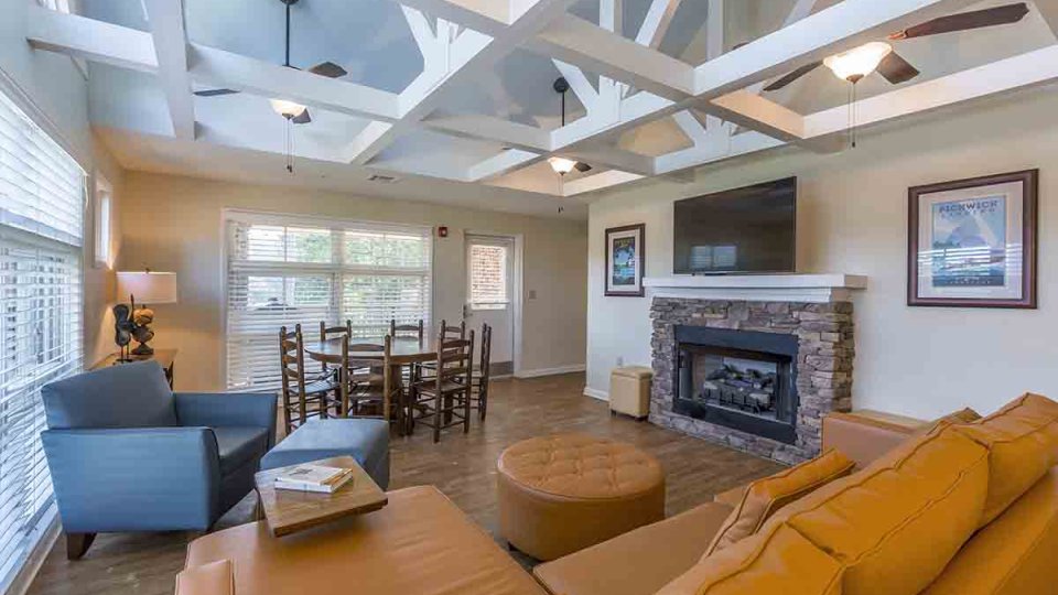 Living room with couch, a cushioned chair, foot rests, dining table, chairs, ceiling fans, overhead lighting, windows, a lamp, a television above the fireplace, and a door that leads outside