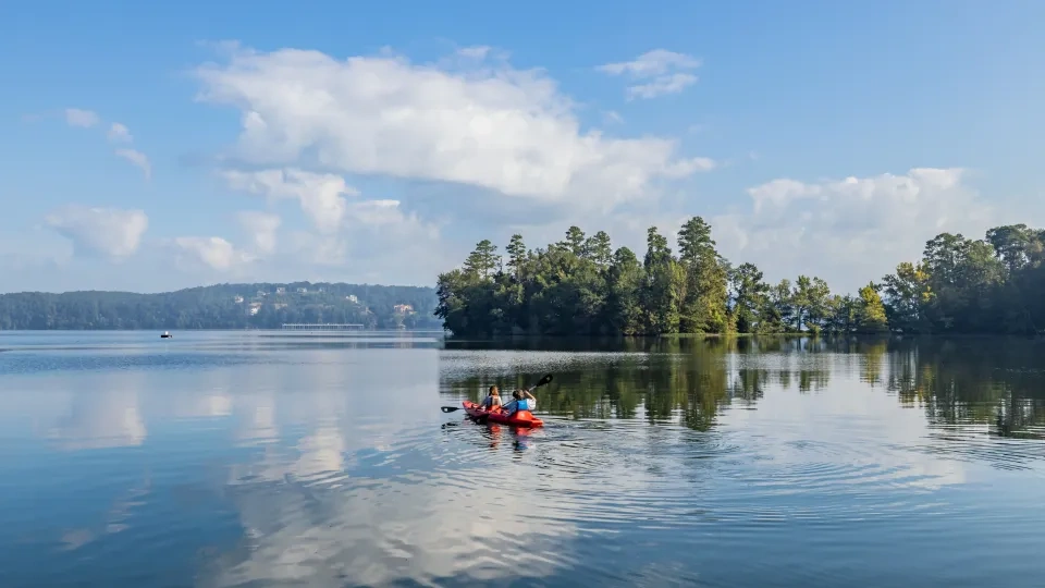Kayakers on calm lake, forested island in background under a bright blue sky.