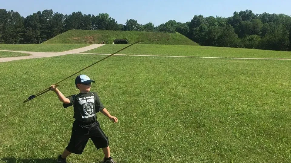Boy with cap throwing spear in grassy field, sunny day.