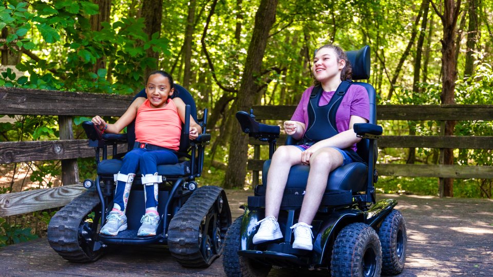 Two smiling kids in wheelchairs on a wooden path in a lush green forest.
