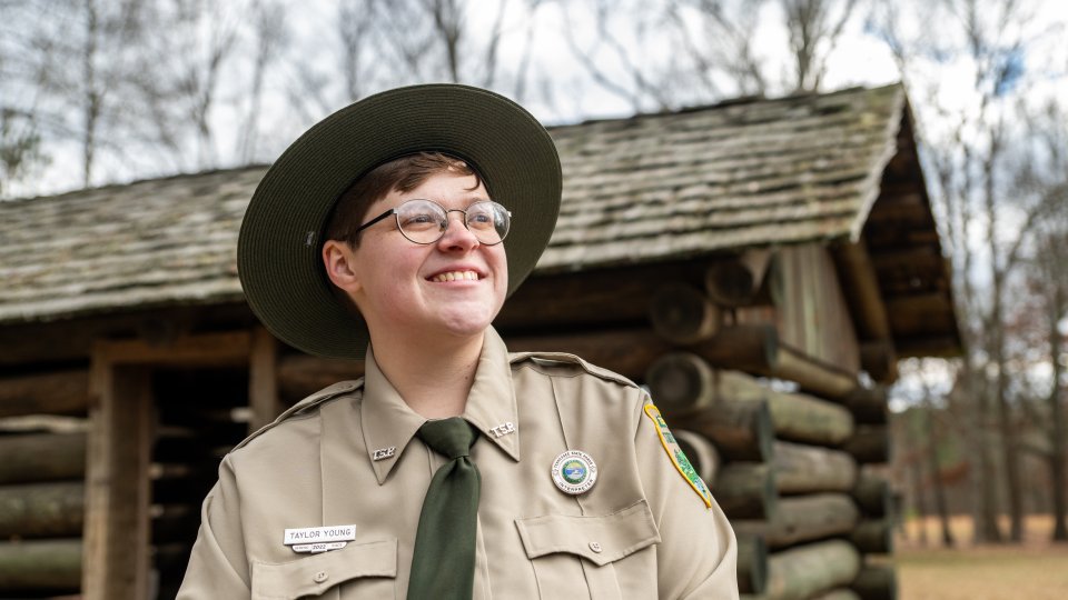 Park ranger in uniform smiles confidently, standing in front of a rustic log cabin, surrounded by leafless trees under a cloudy sky.