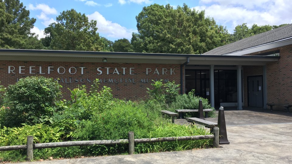 Entrance to Reelfoot State Park Visitor Center. The brick building is surrounded by lush greenery and a wooden fence under a partly cloudy sky.