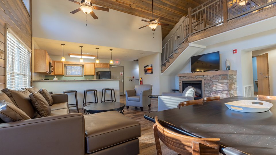 Spacious living room with sofa, kitchen, dining table, stairs, and a stone fireplace.