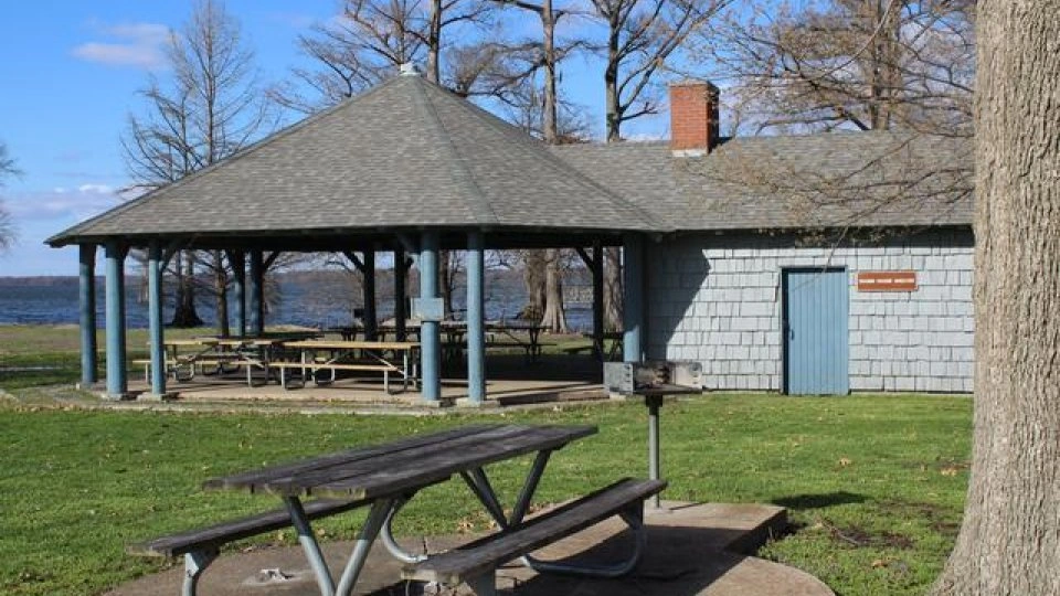 Park pavilion with picnic tables near a lake, surrounded by trees.