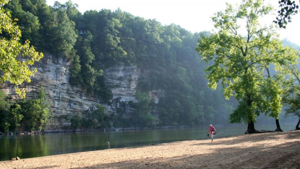 A serene riverside scene with a sandy beach, lush green trees, and towering cliffs. A person in red strolls leisurely, conveying tranquility and natural beauty.