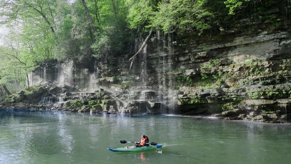 A person kayaking on a calm river near a mossy, rocky cliff with cascading waterfalls. Surrounding lush green foliage creates a serene atmosphere.