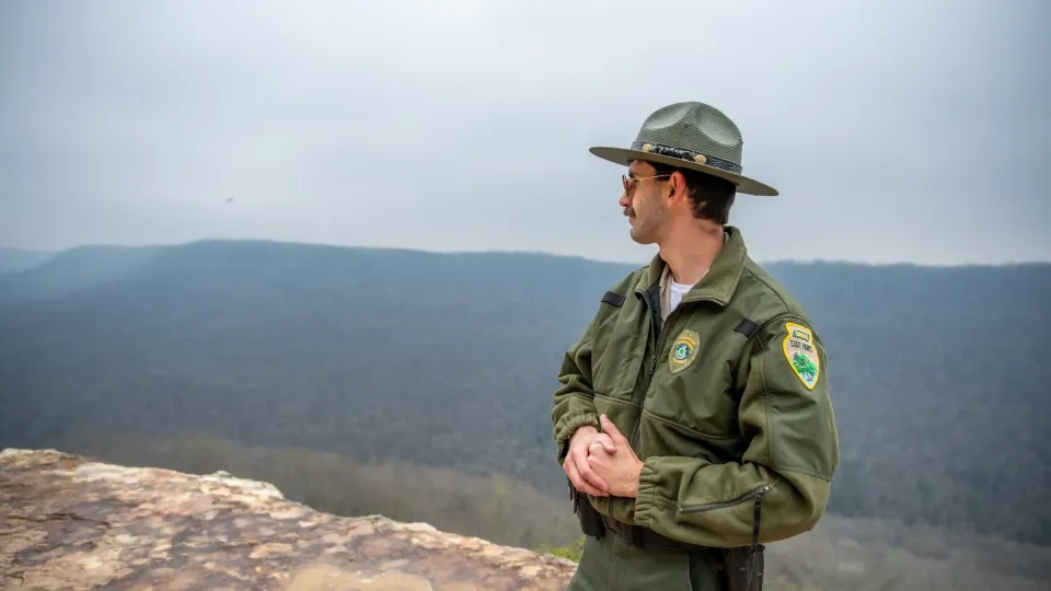 A park ranger stands on a rocky cliff, gazing at the misty, forested mountains. He's wearing a green uniform and hat, exuding a calm, vigilant demeanor.