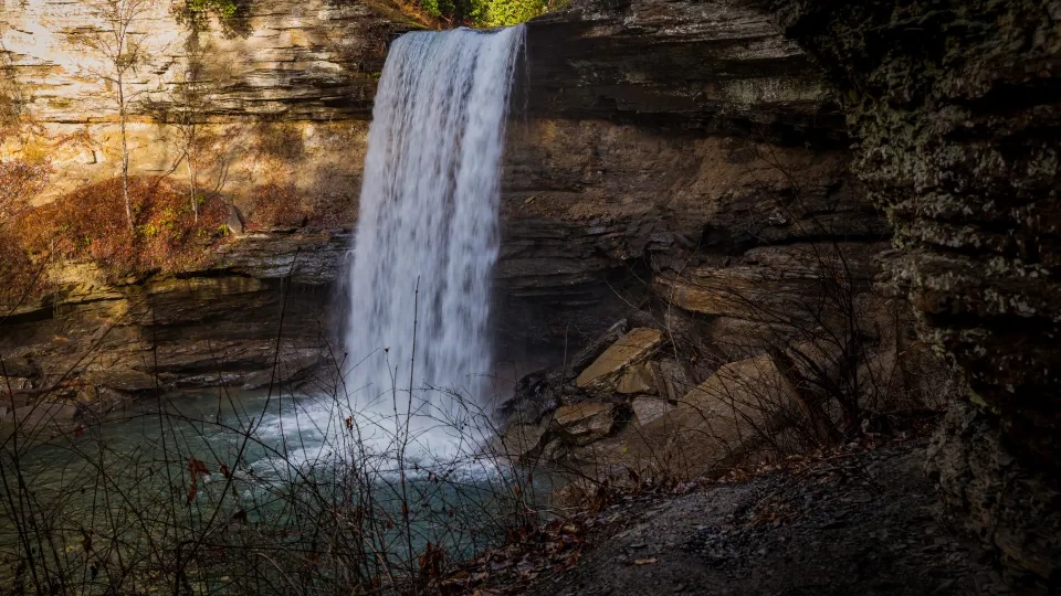 Waterfall cascading over rocky cliff into a turquoise pool, surrounded by dry branches.