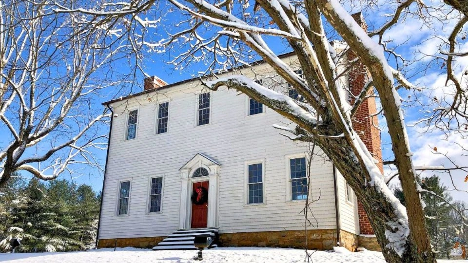 Historic white house with a red door and wreath, surrounded by snow-covered trees, under a bright blue sky. Wintery, peaceful atmosphere.