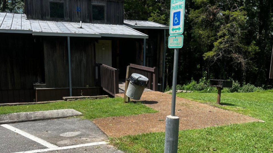 A rustic wooden building with a wheelchair-accessible sign in front, next to a sloped trash can.