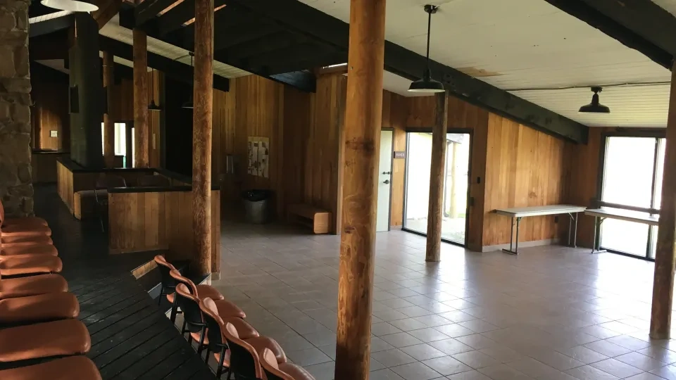 Spacious room with wooden walls, exposed beams, and large windows. It features a row of orange chairs and a long table, evoking a rustic, serene atmosphere.