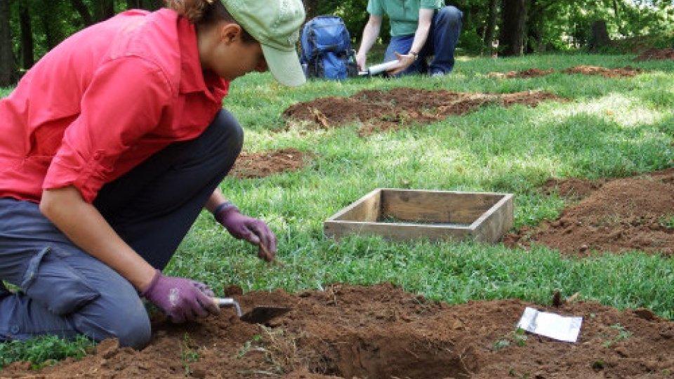 A person in a red jacket digs in a grassy area, surrounded by shallow pits. Another person writes notes in the background. The scene is calm and focused.
