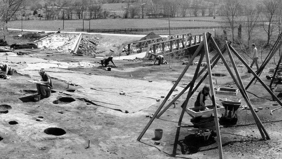 Archaeological dig site in a rural area, featuring workers examining the ground and using sieve stands. Large holes and tools are visible, with trees and fields in the background.