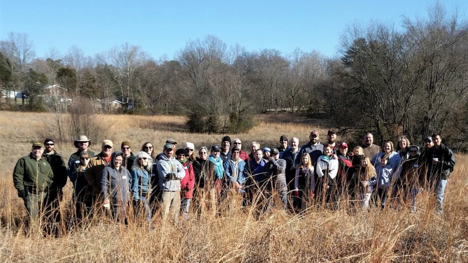 A diverse group of people stands in a field of tall, golden grass under a clear blue sky. They appear cheerful, suggesting a nature walk or group outing.