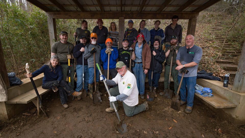 A group of 16 people, smiling and holding tools, poses under a wooden shelter in a wooded area. The mood is upbeat and collaborative.
