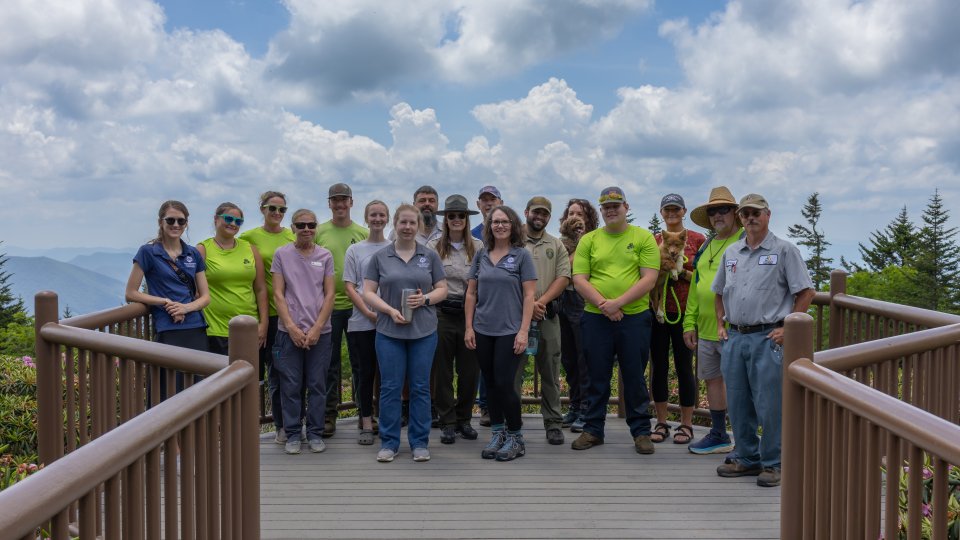 A diverse group of 15 people stands on a wooden deck, smiling in bright sunlight. Some wear vibrant green shirts. A scenic view of mountains and lush trees is behind them under a partly cloudy sky.