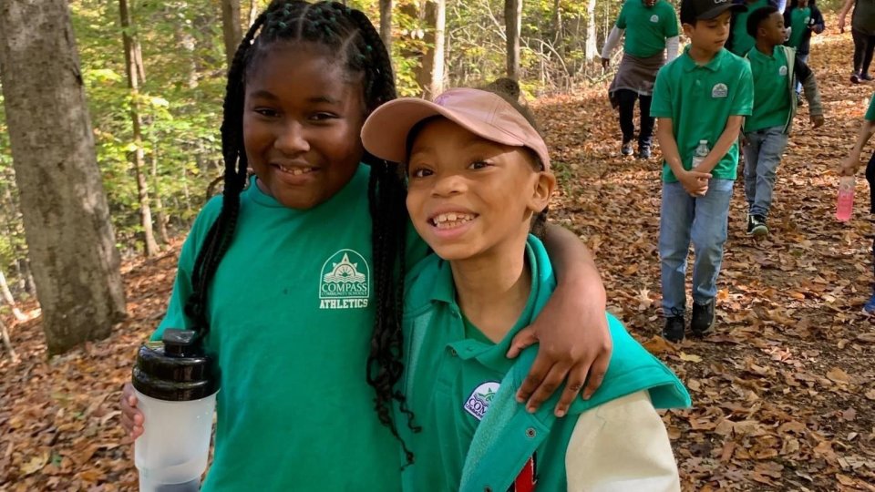 Young girls hugging each other in the forest in front of other kids wearing green in a forest