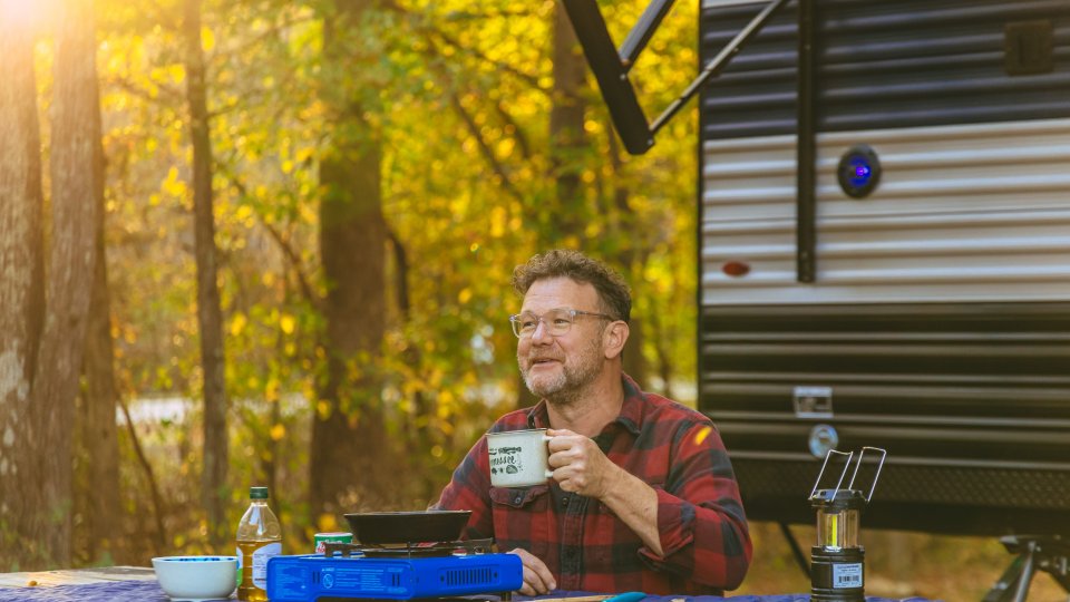 A man sitting at a picnic table in front a camper enjoying a cup of coffee in the morning. 