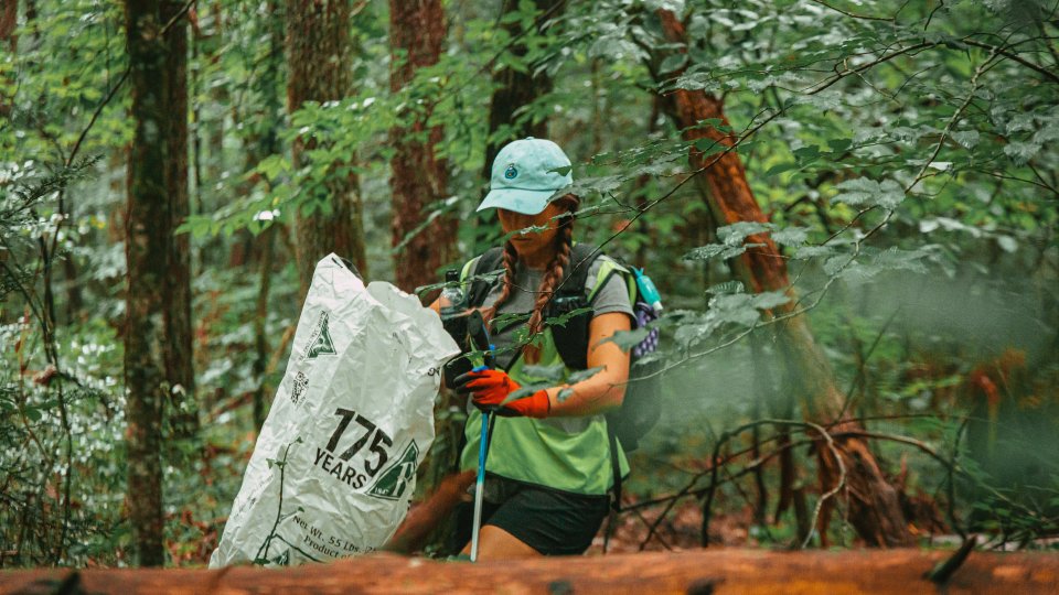 A person in a green shirt and cap, wearing orange gloves, collects trash in a forest with a large white bag. 
