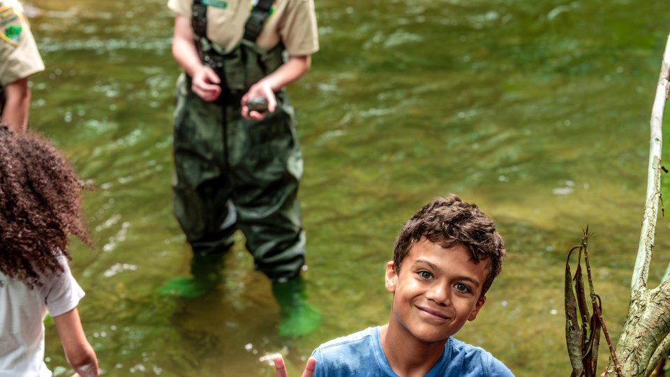 A smiling boy in a blue shirt holds a peace sign beside a stream. A park ranger and another child in shorts wade nearby. The scene is lively and joyful.
