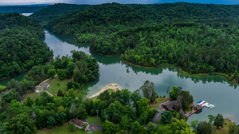 Aerial view of a lush green landscape with a winding river. Trees densely cover hills under a cloudy sky, and small buildings are visible beside the river. Peaceful and serene atmosphere.