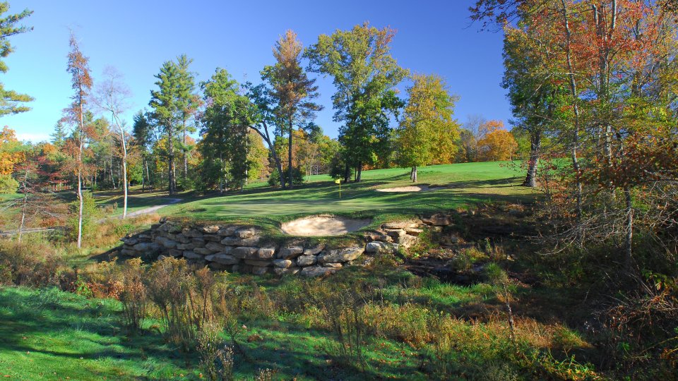 A lush, green golf course is surrounded by autumn trees under a clear blue sky. A sand bunker and rocky edge are in the foreground, creating a serene atmosphere.