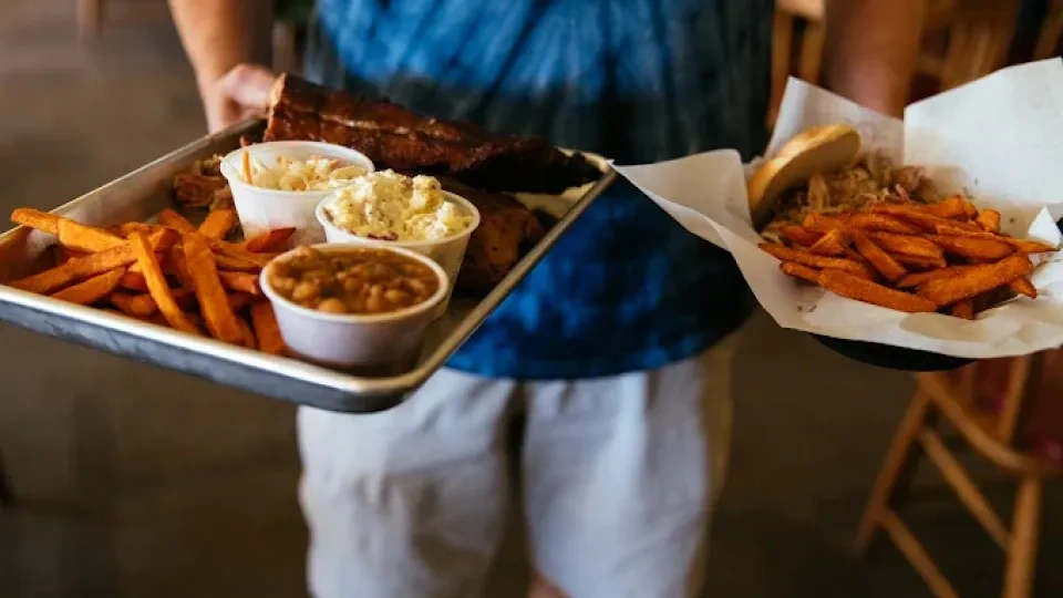 Person holding two trays filled with food, including sweet potato fries, beans, coleslaw, potato salad, and a sandwich, creating a casual dining atmosphere.