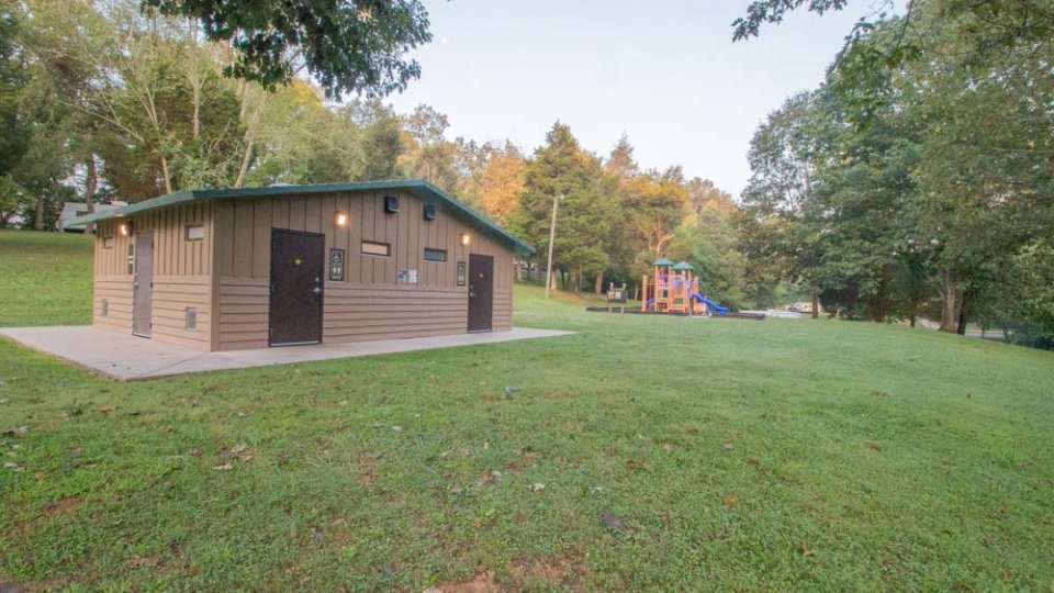A small brown park building with dark doors sits on a grassy field surrounded by trees. In the background, a colorful playground is visible, evoking a serene, inviting atmosphere.