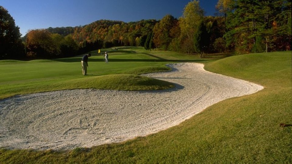 Sunny golf course with two golfers putting near the hole, surrounded by lush green fairways and a sandy bunker. Autumn trees create a serene backdrop.