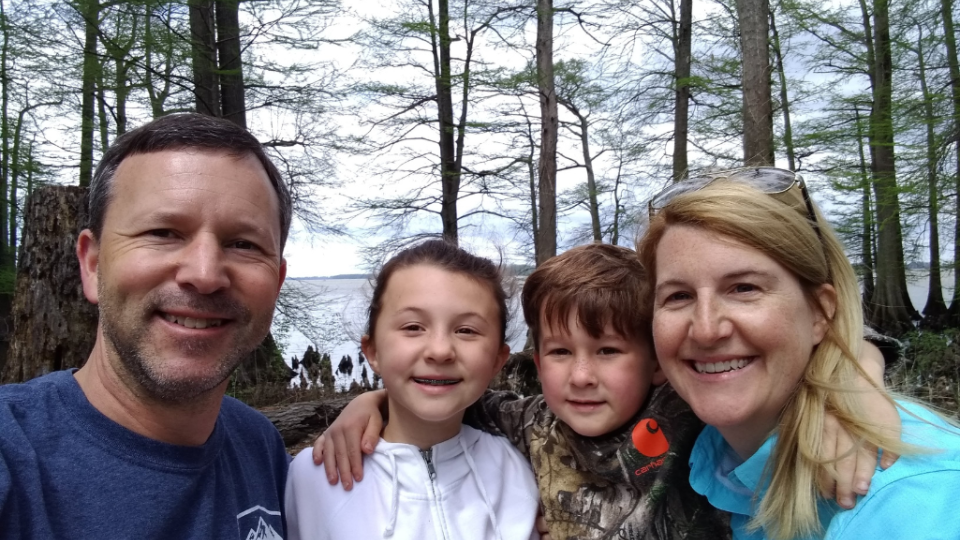 A family of four smiles outdoors in a forest setting, with tall trees and a cloudy sky. The mood is joyful and relaxed, reflecting a sense of togetherness.