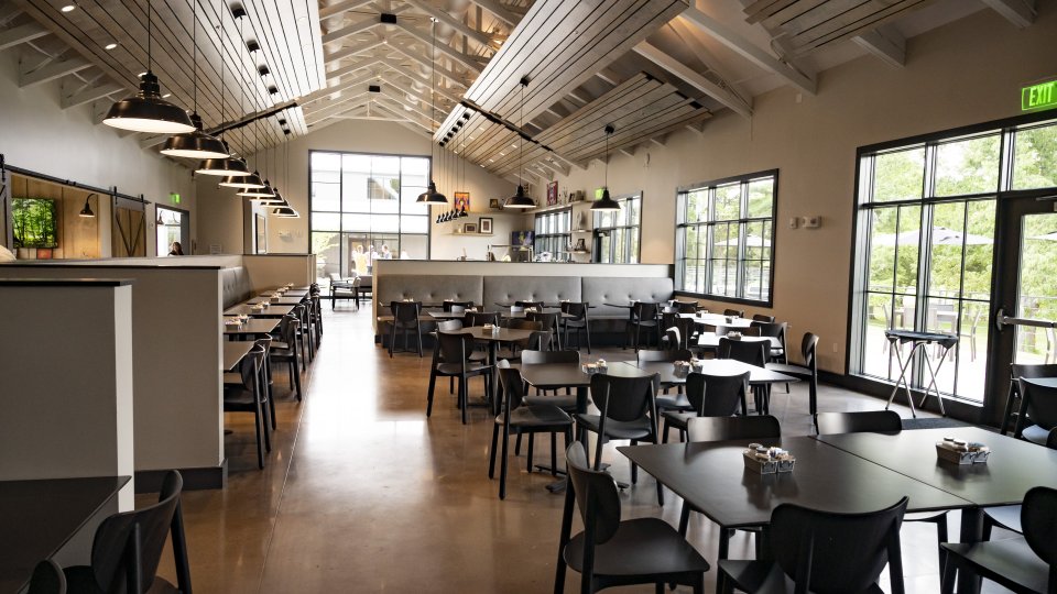 Spacious restaurant interior with high ceilings, modern black tables, and chairs. Large windows let in natural light, creating a calm, inviting atmosphere.