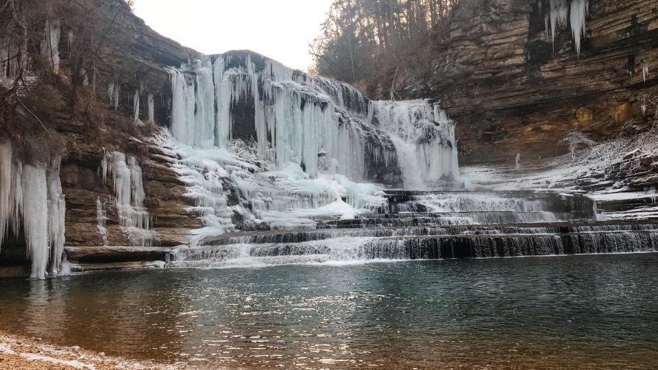A frozen waterfall covered in icicles cascades over layered rock formations into a calm pool. The scene is serene and evokes a sense of winter stillness.