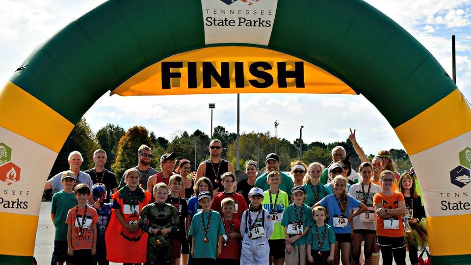 A diverse group of smiling participants stands under a green and yellow "Finish" arch at a Tennessee State Parks event. The atmosphere is cheerful and celebratory.
