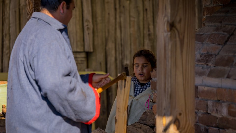 Man in gray coat shows a young girl how to chop wood with an axe. They're in a rustic setting with wooden and brick elements. The tone is instructional.