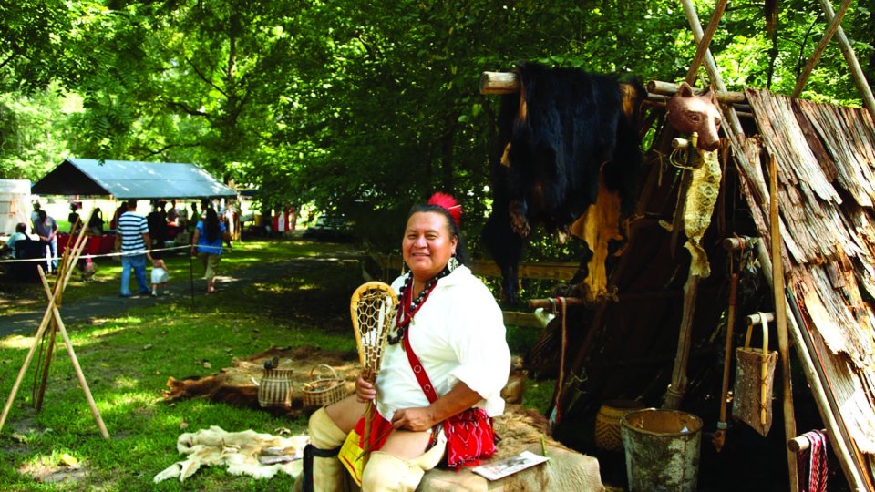 Person in traditional attire sits by rustic shelter with forest and small gathering nearby.