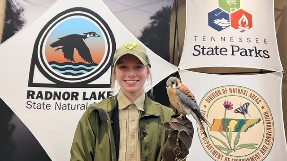 Smiling park ranger holding a small falcon, standing before state park banners.