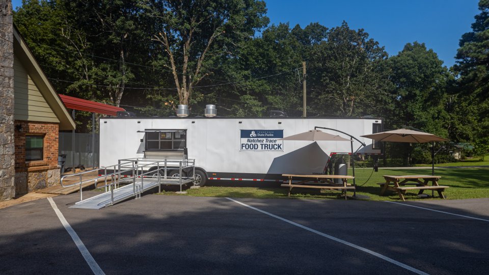 A white food truck in a park, shaded by tall trees and a clear blue sky. Nearby are picnic tables with umbrellas, creating a relaxed outdoor eating area.