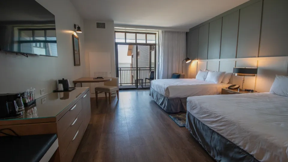 Two queen size beds in a bedroom with a television, dresser, desk, balcony, and lighting