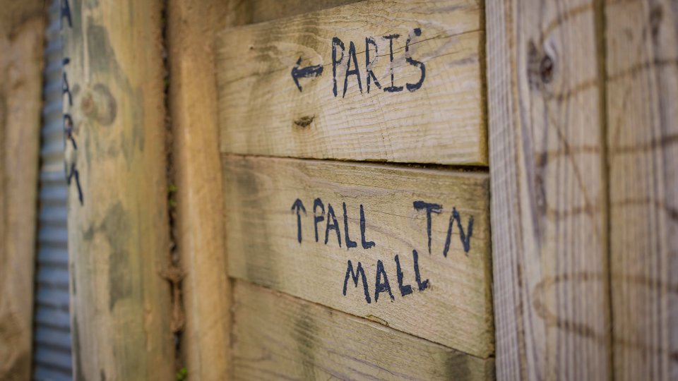 Wooden wall with handwritten directional signs in black ink. One arrow points left to "Paris," another points up to "Pall TN Mall." Rustic and casual vibe.