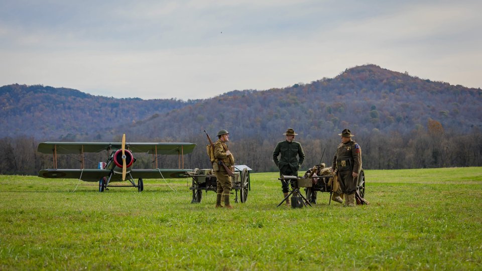 Three people in historical military uniforms stand on a grassy field beside a vintage biplane. A mountain range is visible in the background, under a cloudy sky.