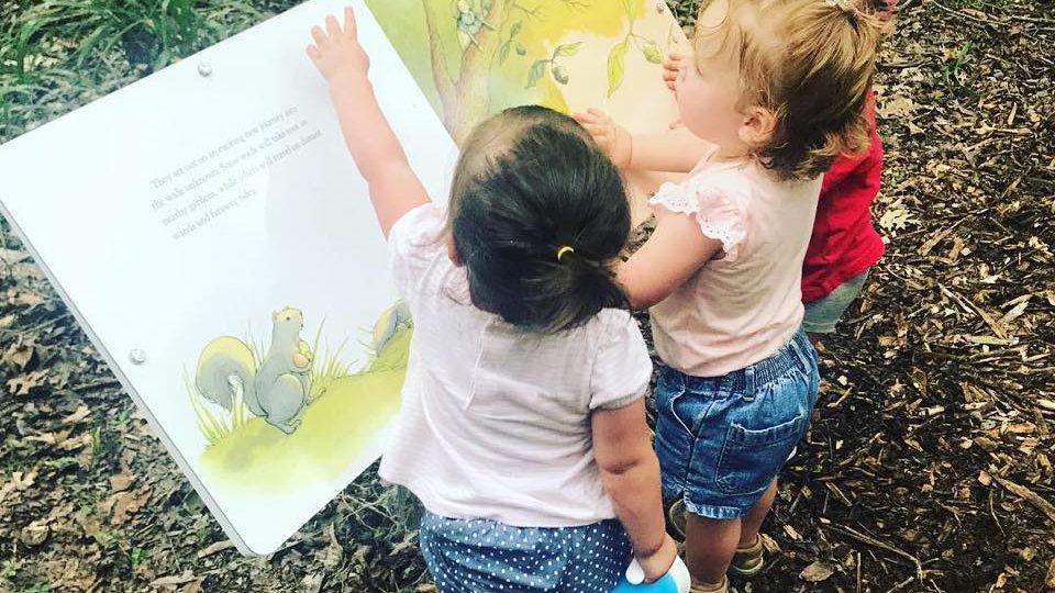 3 children hovering over the storybook trail signage in the woods on a trail.