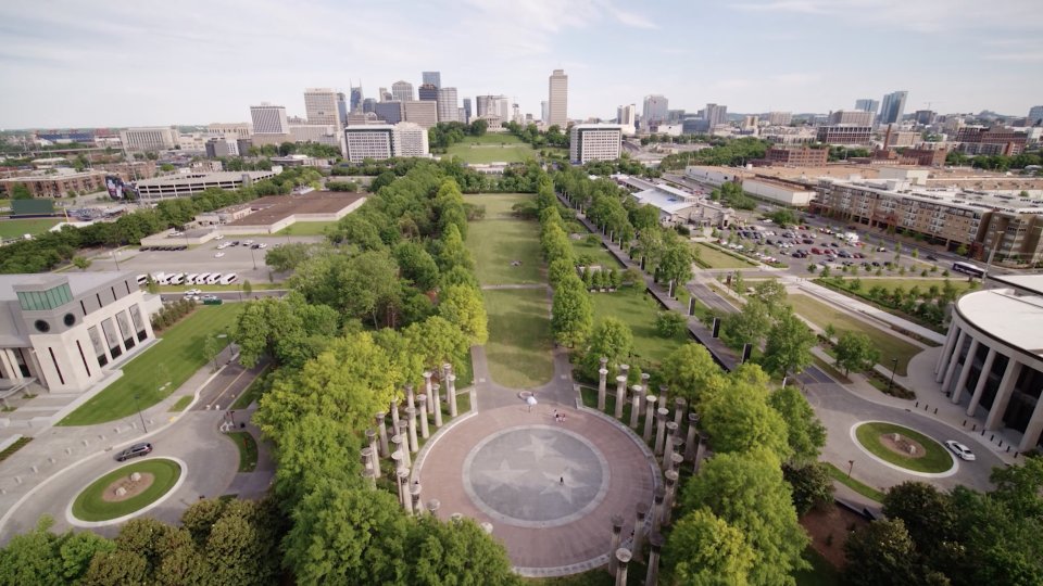 Aerial view of a city park with a circular plaza featuring a star pattern. Tree-lined paths extend towards a city skyline under a clear blue sky.