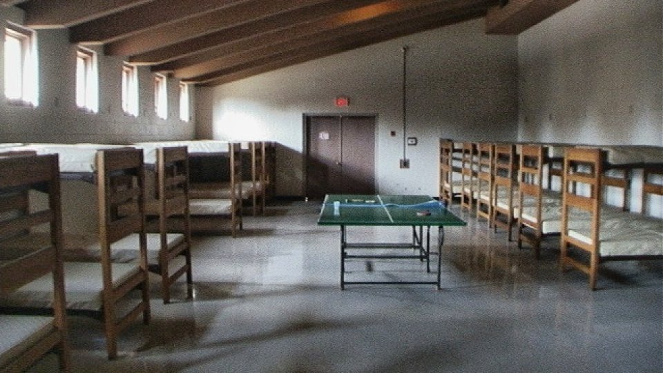 A dimly lit dormitory with empty bunk beds lining the walls, a ping-pong table centered on the polished floor, and small windows casting soft light.