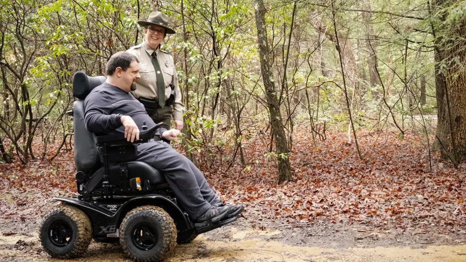 Man in wheelchair and park ranger smiling on forest trail.