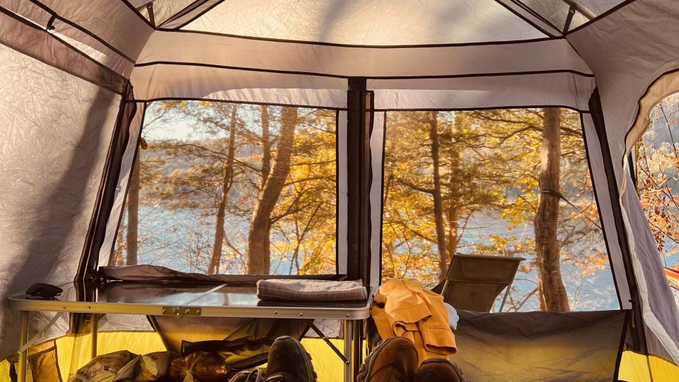 A person relaxes in a hammock near a campfire, surrounded by autumn trees. A lake reflects the warm sunset, creating a serene and peaceful atmosphere.